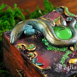 Può includere: Una scatola di legno con un design di lucertola verde e oro. La scatola è decorata con perline e una gemma verde. Il testo "Fogliaviola Fantasy Design" è sulla scatola.