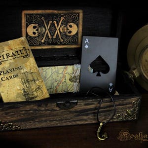 May include: A wooden box with a brass latch and ornate trim. Inside the box are a deck of playing cards with a pirate ship design, a map, and a black metal ace of spades bottle opener. The box is sitting on a dark surface.
