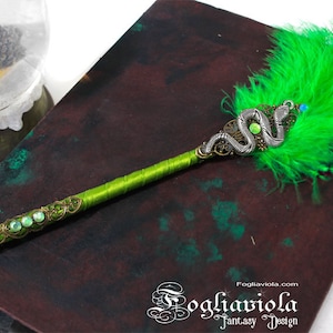 Può includere: Una penna stilografica verde con un designo a serpente argentato e pietre preziose verdi. La penna è appoggiata su una superficie verde scuro e marrone.