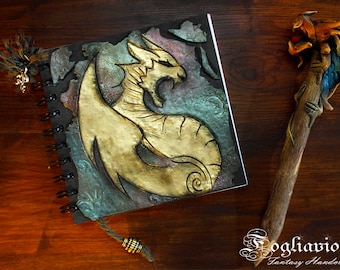 May include: A spiral bound journal with a gold dragon design on the cover. The dragon is sculpted and has a textured, metallic finish. The journal is on a wooden surface with a decorative wooden wand next to it.