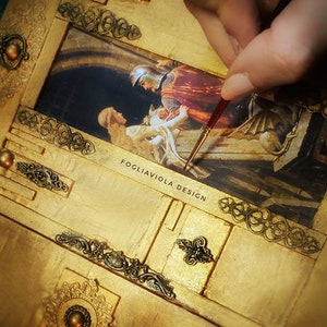 May include: A gold-toned wooden box with intricate metal accents and a painted scene of a knight rescuing a woman. The box is being painted with a fine brush. The text "FOGLIAVIOLA DESIGN" is visible on the box.