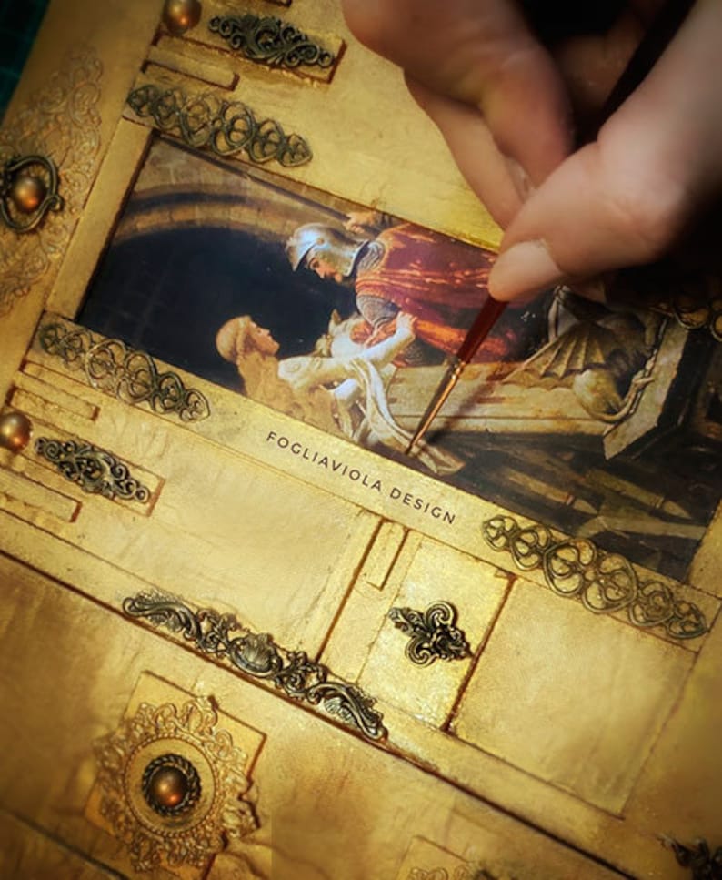 May include: A gold-toned wooden box with intricate metal accents and a painted scene of a knight rescuing a woman. The box is being painted with a fine brush. The text "FOGLIAVIOLA DESIGN" is visible on the box.