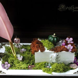 Può includere: Un vassoio bianco decorativo con un forziere in miniatura, muschio, fiori e una penna rosa. Il vassoio è adornato con angoli in bronzo e un pomello verde a forma di zucca. La scena evoca un tema fantasy.