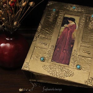 May include: A gold-colored book with a decorative design and turquoise gemstones. The cover features a vintage illustration of a woman in a long red dress holding a small orb.