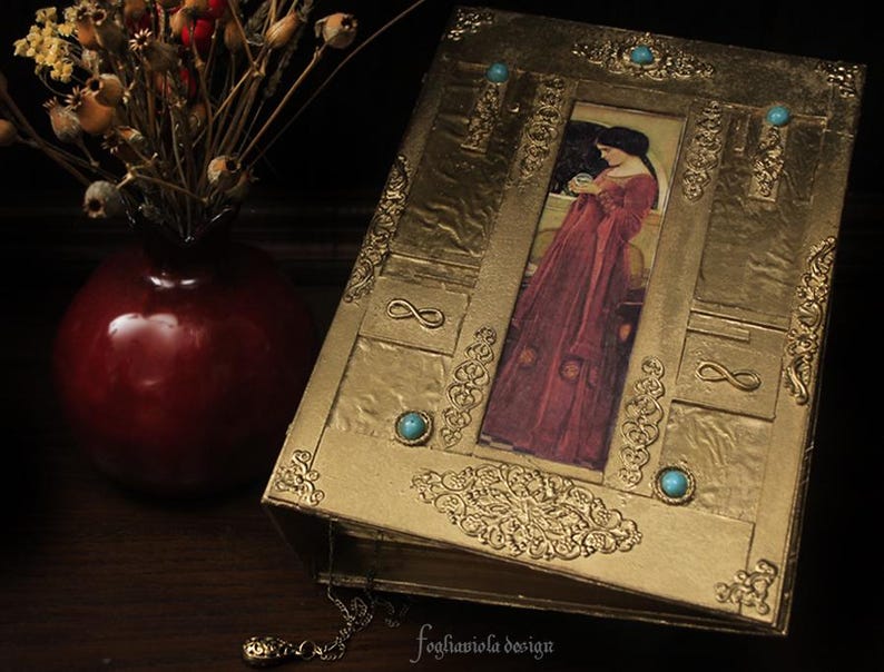 May include: A gold-colored book with a decorative design and turquoise gemstones. The cover features a vintage illustration of a woman in a long red dress holding a small orb.