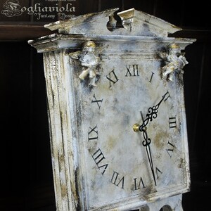 Puede incluir: Reloj blanco y dorado desgastado con números romanos y detalles de querubines. La esfera del reloj es blanca con agujas negras y números romanos. El reloj está sobre un soporte.