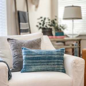 K&ouml;nnte beinhalten: Ein wei&szlig;er Sessel mit einem blauen und grauen Kissen mit Muster und einer blauen Wolldecke. Der Sessel steht in einem Wohnzimmer mit einem Beistelltisch und einer Lampe im Hintergrund.