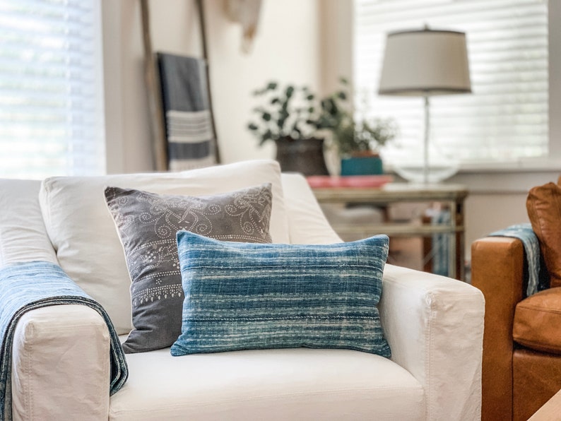 K&ouml;nnte beinhalten: Ein wei&szlig;er Sessel mit einem blauen und grauen Kissen mit Muster und einer blauen Wolldecke. Der Sessel steht in einem Wohnzimmer mit einem Beistelltisch und einer Lampe im Hintergrund.