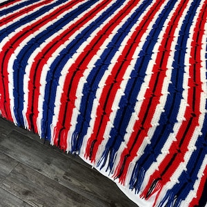 May include: A handmade, fringed blanket with a striped pattern in red, white, and blue. The blanket is crocheted with a textured stitch, and the fringe is at the bottom edge. The blanket is draped over a white sheet.