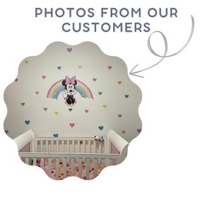 Pode incluir: Um ber&ccedil;o branco com um len&ccedil;ol estampado com flores e uma parede branca com um arco-&iacute;ris e um adesivo de Minnie Mouse. A parede &eacute; decorada com cora&ccedil;&otilde;es coloridos.