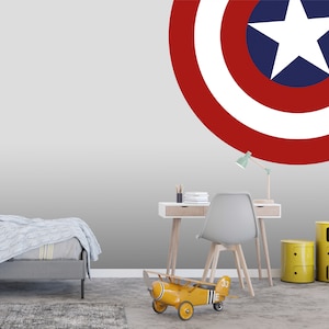 May include: A gray bedroom with a white wall featuring a red, white, and blue Captain America shield decal. A gray bed with a blue knitted blanket is in the foreground. A yellow toy airplane is on the floor. A desk with a chair and a lamp is in the background.