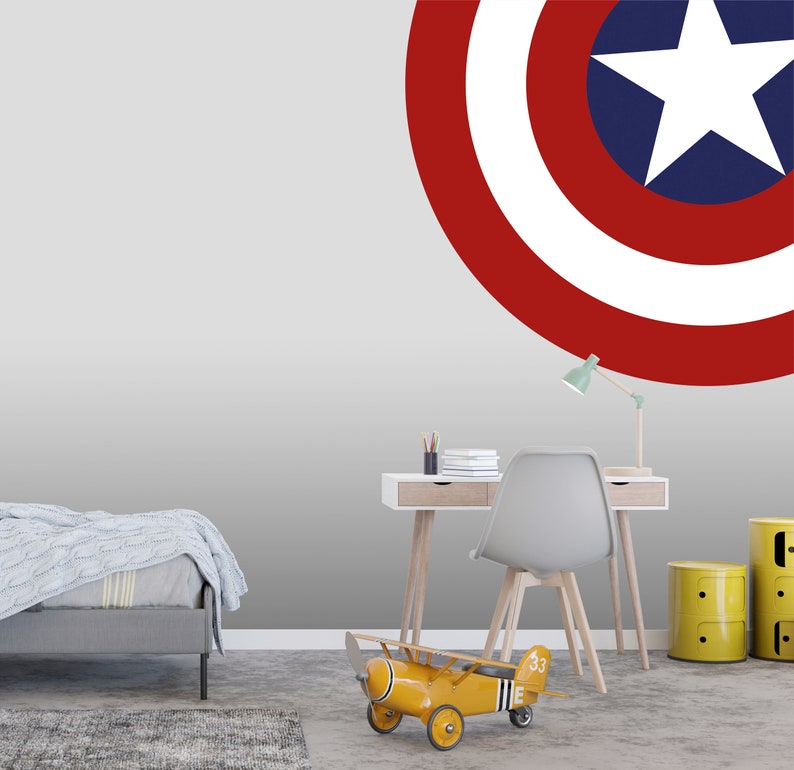 May include: A gray bedroom with a white wall featuring a red, white, and blue Captain America shield decal. A gray bed with a blue knitted blanket is in the foreground. A yellow toy airplane is on the floor. A desk with a chair and a lamp is in the background.