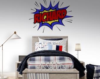 May include: A bedroom scene featuring a bed with a gray wooden frame, adorned with a blue and white plaid comforter and decorative pillows. Above the bed, a wall decal displays the name "RICHARD" in a comic book style. A floor lamp and a bedside lamp are also visible.