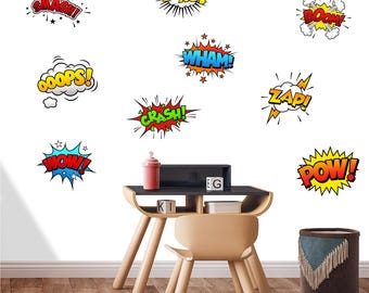 May include: A collection of colorful comic book-style sound effect wall decals, including "SMASH!", "BANG!", "BOOM!", "WHAM!", "OOOPS!", "ZAP!", "CRASH!", and "POW!". A small wooden desk and chair are in the foreground, with a red container and a decorative basket.