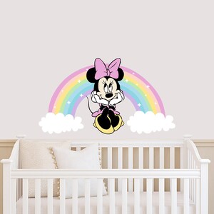 Pode incluir: Um ber&ccedil;o branco com um adesivo de parede Minnie Mouse rosa e arco-&iacute;ris. O adesivo apresenta Minnie Mouse sentada em um arco-&iacute;ris com nuvens de cada lado.