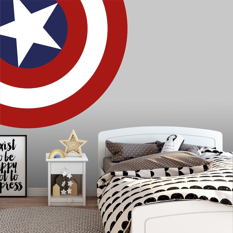 May include: A white bed with a black and white patterned duvet cover and a red, white, and blue Captain America shield wall decal. The bed is in a room with a white nightstand and a framed print with the text "Exist to be happy, not to impress".