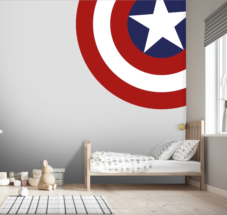 May include: A white, red, and blue Captain America shield decal on a gray wall in a child's bedroom. The decal is in the shape of a circle and has a white star in the center. The room also features a wooden bed with white bedding and a wooden nightstand.
