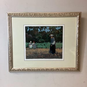 May include: Framed art print featuring a pastoral scene with figures in a field. The artwork depicts three women, one standing and two seated, with a backdrop of trees and a blue sky. The frame is silver-colored with ornate detailing.