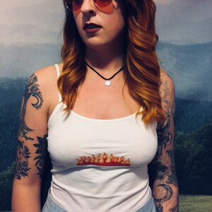 May include: A white tank top with a red and orange flame graphic. The model is wearing round, tinted sunglasses and a black choker necklace.
