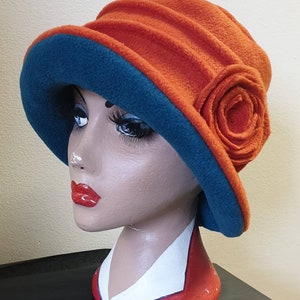 May include: A rust-colored fleece hat with a teal blue lining and a large rust-colored fabric flower.