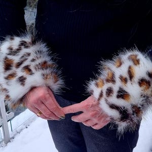 Puede incluir: Un par de calentadores de brazo de piel sintética blanca con estampado de leopardo marrón y negro. Los calentadores de brazo se llevan en los brazos de una persona.