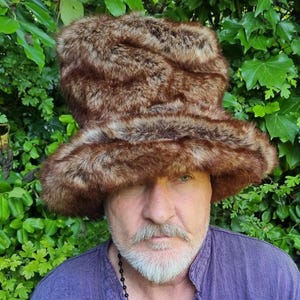 May include: A tall, cylindrical hat made of brown and tan faux fur with a wide, flared brim. The hat features a dense, textured pile with variegated colour patterns throughout, creating a thick, voluminous appearance that covers the top of the head.