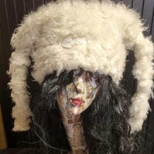 Puede incluir: Sombrero blanco y peludo con orejas largas y caídas. El sombrero tiene una etiqueta blanca con el texto "Off My Head!" en él.