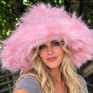 May include: A wide-brimmed bucket hat made of long, shaggy, bright pink faux fur material. The hat features a deep crown and a floppy, oversized brim that frames the face with its dense, textured, and voluminous synthetic fiber construction.