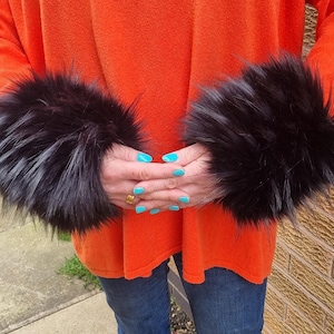 Puede incluir: Un par de calentadores de brazo de piel sintética negra. Los calentadores de brazo se llevan sobre un jersey naranja brillante y unos vaqueros azules.