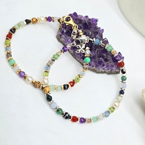May include: A multi-colored beaded bracelet with a gold clasp. The bracelet features a variety of gemstones, including amethyst, aquamarine, and emerald. The bracelet is displayed on a purple amethyst cluster.