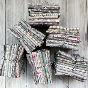 May include: Several bundles of gray fabric, each tied with black and white twine. The fabric has various patterns, including floral, geometric, and striped designs. The color palette is primarily gray, with accents of white, pink, and teal. The bundles are stacked on a light wood surface.