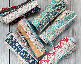 May include: Rolled fabric bundles featuring floral and geometric patterns, tied with twine. The fabric displays a range of colours including blue, red, green, and brown. Suitable for quilting or craft projects, the fabric appears to be cotton.