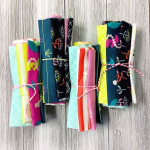 May include: Four bundles of colorful fabric rolls tied with twine. The fabric features floral, geometric, and striped patterns in shades of teal, pink, yellow, and black. The bundles are arranged on a white wooden surface.