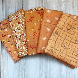 May include: A collection of folded fabric pieces in shades of orange and gold, featuring various floral and geometric patterns. The fabrics display designs with red, white, and green flowers, and dotted and patterned details.