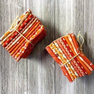 May include: Two bundles of orange and cream-colored fabric strips, tied with yellow twine. The fabric features various patterns, including floral and geometric designs. The bundles are arranged on a light gray wooden surface.