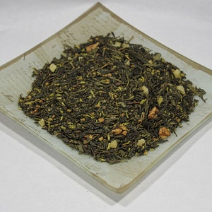 May include: A close-up of a light green tea blend with pieces of dried fruit and nuts. The tea is in a square, light blue and white ceramic dish.