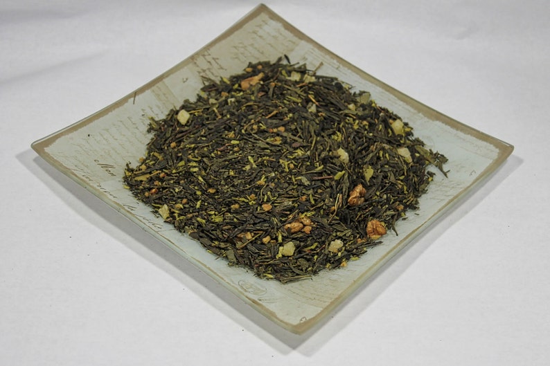 May include: A close-up of a light green tea blend with pieces of dried fruit and nuts. The tea is in a square, light blue and white ceramic dish.