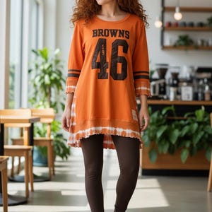 May include: An orange long-sleeve tunic featuring the text BROWNS and the number 46 in dark brown, with dark brown stripes on the sleeves and a white tie-dye hem. The tunic is paired with dark brown leggings and bright orange slip-on clogs.