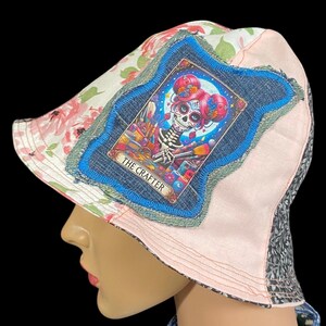 May include: A patchwork bucket hat featuring floral, pink, and denim fabrics. The hat has a decorative patch with a Day of the Dead-style illustration and the text "THE CRAFTER". The hat is designed with a brim and a rounded crown.