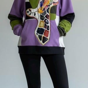 Puede incluir: Una sudadera con capucha morada con un diseño de jirafa. La sudadera tiene un bolsillo de canguro, detalles en blanco y negro y mangas verdes y negras. La persona lleva leggings negros y zuecos rojos.