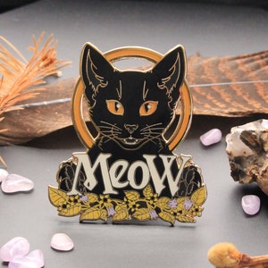 Peut inclure: Épinglette en émail noir représentant un chat noir aux yeux jaunes et le mot "MEOW" en lettres blanches. Le chat est entouré de détails floraux dorés.