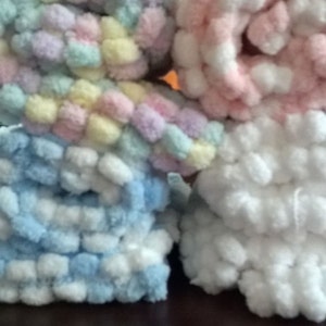May include: Three skeins of yarn in different colors: pastel rainbow, pink and white, and white and light blue. The yarn is made of soft, fluffy pom-poms.