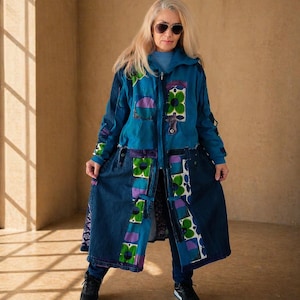 Op de afbeelding: Een lange patchwork denim jas met een teal bovenstuk en een donkerblauw onderstuk. De jas heeft bloemen- en geometrische patronen in groen, paars en wit. De jas wordt gedragen met een donkerblauwe jeans en zwarte sneakers.
