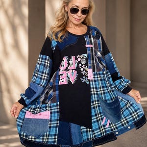 Op de afbeelding: Een tuniek met lange mouwen in patchworkstijl, voorzien van een zwart middenpaneel met de roze geborduurde tekst LOVE en bloemmotieven. Het kledingstuk is opgebouwd uit diverse blauwe flanellen ruitjes- en denimstoffen, inclusief meerdere zakken en een gerafelde zoom.