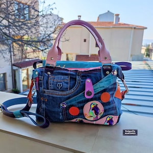 May include: A denim handbag with a pink handle and strap, featuring a bird illustration on a pink patch. The bag has a blue strap and orange and teal accents. The text "Designed by MARTINELLI" is visible.