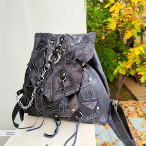 May include: A black backpack featuring a mix of textured fabric and faux leather panels with silver-tone metal studs, rings, and a chain detail. The front flap includes geometric overlays and hanging leather cords. Text reads: Designed by MARTINELI.