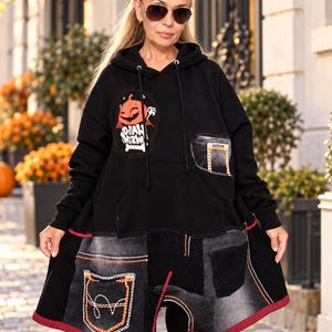 Puede incluir: Una sudadera negra con capucha que presenta un gráfico de una calabaza naranja con el texto "HALLOWEEN" y un hueso. La parte inferior está confeccionada con paneles de mezclilla gris oscuro con costuras naranjas y varios bolsillos.