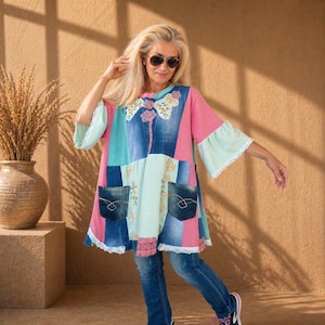 Op de afbeelding: Een patchwork tuniek met panelen van blauwe denim, roze stof en lichtblauw materiaal. Het kledingstuk is voorzien van twee voorzakken, bloemenborduursel op de borst en witte kanten randen langs de zoom en de mouwen.