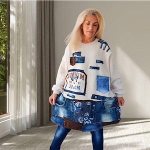 Pode incluir: Uma camisola de manga comprida em branco sujo com um design de retalhos de tecido de ganga, um bolso castanho e uma impressão gráfica central da Minnie Mouse com o texto "MINNIE MOUSE 28". A parte inferior é uma sobreposição de saia de ganga.