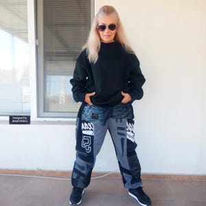 Può includere: Una persona indossa una felpa nera e pantaloni in denim patchwork. I pantaloni presentano pannelli grigi e neri con testo e grafica bianchi. Le scarpe da ginnastica nere completano l'outfit. Il testo "Designed by MARTINELLI" è visibile.